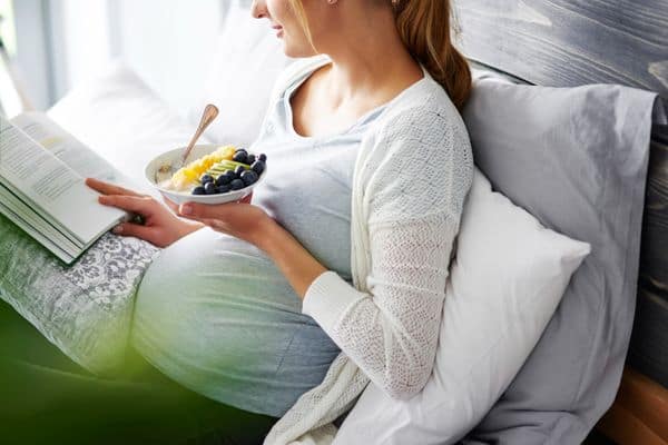 אישה בהריון קוראת ספר ואוכלת קערת פירות